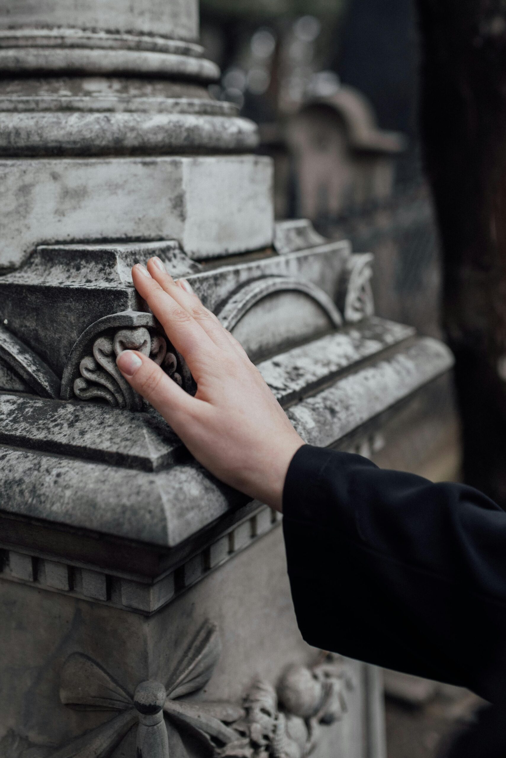Close-up of a hand touching an ornate stone structure in a cemetery, conveying remembrance.
